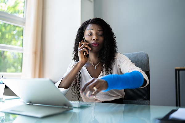 Woman with blue arm cast talks on the phone while sitting at a laptop computer.