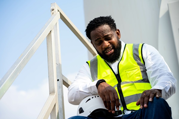 A construction worker with a beard is sitting on a metal staircase, wearing a high-visibility yellow safety vest over a white shirt. He appears to be tired or in discomfort, with a pained expression on his face. His left hand is placed on his knee, and his right hand is resting on a white safety helmet.