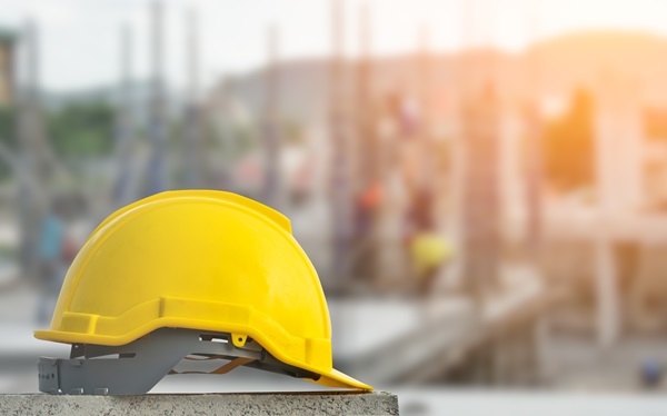 Yellow construction helmet placed on a concrete surface at a building site, with blurred workers and structures in the background under warm sunlight.