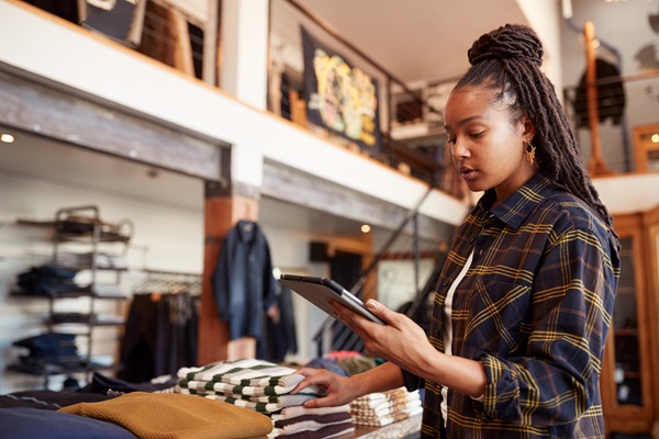 Woman in a retail clothing store using a tablet while organizing folded sweaters on a display table, representing small business operations and workplace settings.