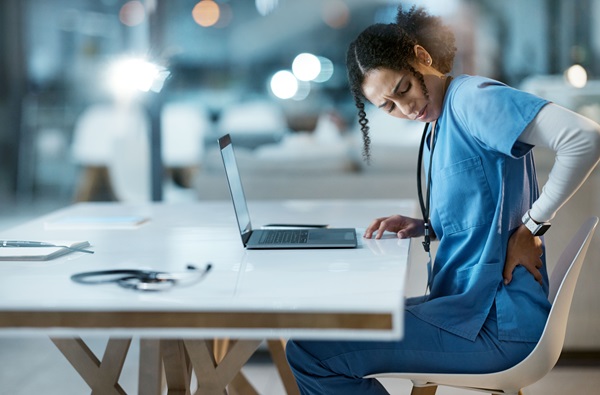 A nurse holds her lower back in pain while sitting at a work station in front of a laptop.