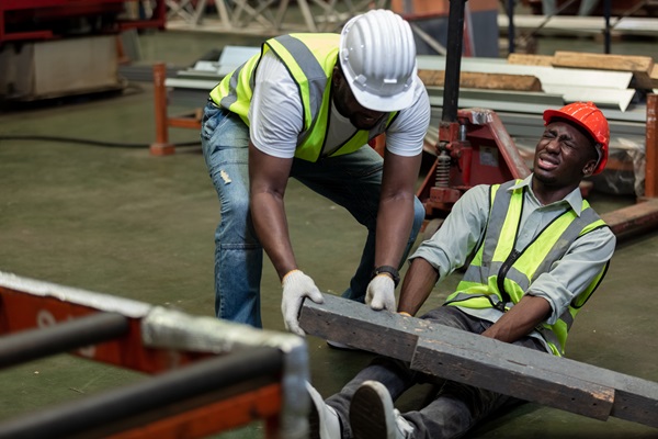 Two construction workers wearing safety vests and hard hats are in an industrial setting. One worker appears injured on the floor, wincing in pain, while the other tries to lift a heavy beam off his leg.