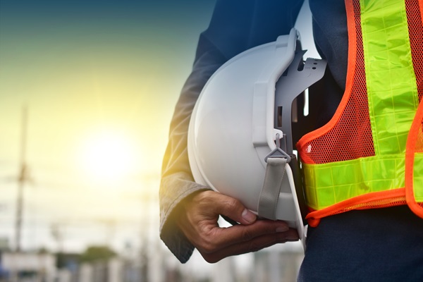A New York City construction worker in a high-visibility orange vest holding a white hard hat tucked under their arm against a bright, sunlit outdoor background.