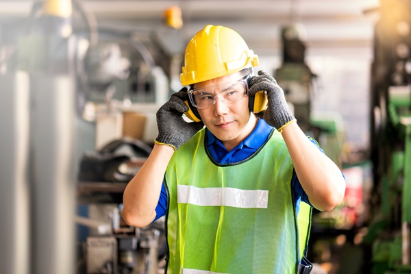 An industrial worker wearing a hard hat and safety vest adjusting protective earmuffs in a loud factory setting, illustrating the occupational hazards that lead to tinnitus and hearing loss disability claims under New York workers' comp.